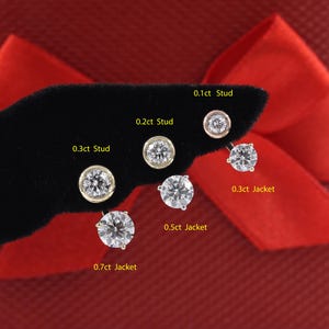 May include: A close-up of ten diamond earrings on a black velvet display. The earrings are arranged in order of size, with the smallest earring on the left and the largest earring on the right. The earrings are labeled with their carat weight and style, such as "0.1ct Stud", "0.2ct Stud", "0.3ct Stud", "0.3ct Jacket", "0.5ct Jacket", and "0.7ct Jacket".