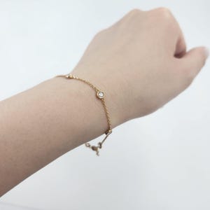 May include: A delicate gold bracelet with several small, round, clear gemstones. The bracelet is on a thin gold chain and is worn on a wrist. The background is a plain white.