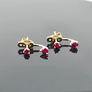 May include: A pair of gold-toned earrings with three red gemstones each. The earrings have a curved design with a post and a small, rectangular backing. The gemstones are round and pear-shaped.