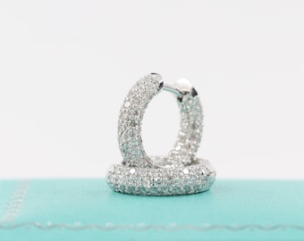 Puede incluir: Un par de pendientes de aro de plata con engaste de diamantes pavé. Los pendientes están sobre un fondo azul claro.