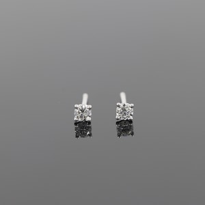 May include: A pair of small, square-shaped stud earrings. Each earring features a single, sparkling, round-cut diamond. The earrings are set on a silver or white gold post, reflecting light. The background is a solid, neutral gray.