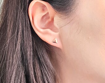 Könnte beinhalten: Ein Paar goldene Dreiecks-Ohrstecker. Die Ohrringe sind klein und schlicht und würden eine großartige Ergänzung für jede Schmucksammlung sein.