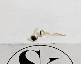 Könnte beinhalten: Ein Paar goldene Ohrstecker mit schwarzen Edelsteinen. Die Ohrringe haben ein schlichtes, elegantes Design mit einem runden schwarzen Stein, der in eine goldene Lünette gefasst ist. Die Stifte sind ebenfalls aus Gold.