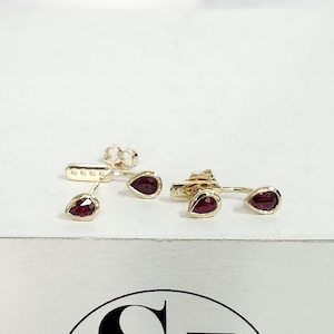 May include: A pair of gold earrings with red teardrop-shaped gemstones. The earrings are designed to be worn in two different ways, with the gemstones either facing forward or backward.