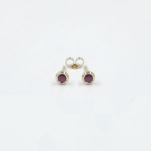 May include: A pair of gold stud earrings with a round, deep red gemstone in each setting. The earrings are displayed with a butterfly earring back on a white background. The gemstones are a vibrant contrast to the gold setting.