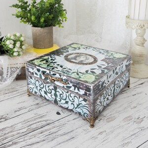 Könnte beinhalten: Dekorative Holzbox mit floralem und Ziegelmuster. Die Box hat ein ovales Porträt, Messingbeschläge und verzierte Eckfüße. Die Farbpalette umfasst Weiß, Grün und Braun.