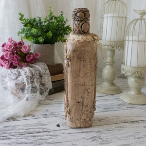 May include: A decorative bottle with a distressed, beige finish and intricate details. The bottle is decorated with lace and has a rustic, vintage look.