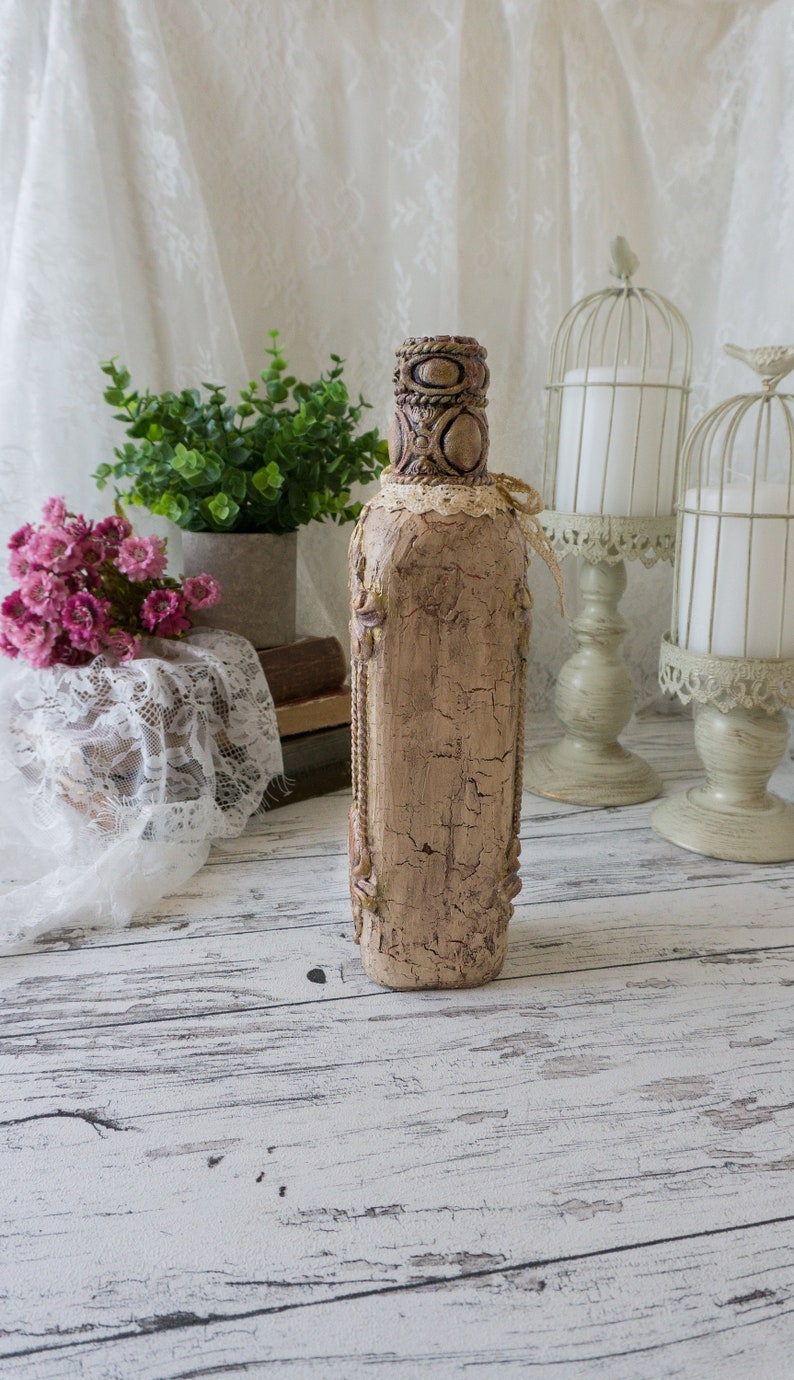 May include: A decorative bottle with a distressed, beige finish and intricate details. The bottle is decorated with lace and has a rustic, vintage look.