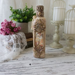 May include: A decorative glass bottle with a beige and brown finish. The bottle has a vintage design with intricate details and a floral pattern. The bottle is sitting on a white wooden surface.