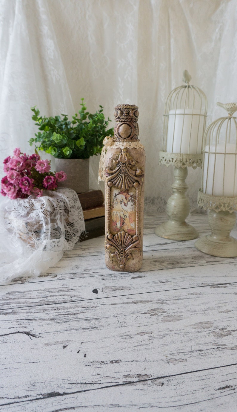 May include: A decorative glass bottle with a beige and brown finish. The bottle has a vintage design with intricate details and a floral pattern. The bottle is sitting on a white wooden surface.