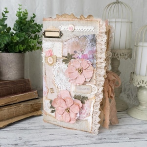 May include: A handmade journal featuring a beige fabric cover decorated with layered lace, paper ephemera, and two large pale pink fabric flowers. The spine is accented with a frayed tan ribbon, and the front cover includes a small metal label frame.