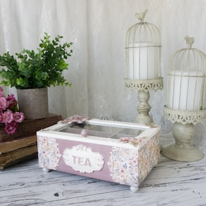 Könnte beinhalten: Eine rechteckige Teebox aus Holz mit altrosa Oberfläche, Glasdeckel und einem zentralen Etikett mit der Aufschrift TEA. Die Box verfügt über floral gemusterte Seitenwände und kleine weiße Füße. Daneben stehen zwei cremefarbene Vogelkäfig-Kerzenhalter.