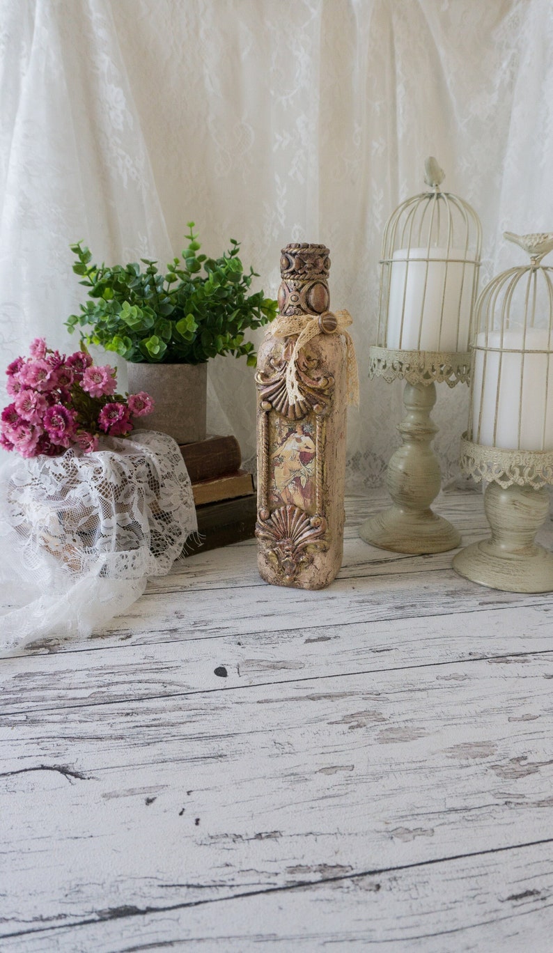 May include: A decorative bottle with a vintage design, featuring a floral pattern and gold accents. The bottle is displayed on a whitewashed wooden surface with two birdcages and a potted plant.