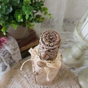 May include: A decorative bottle with a beige and brown finish, featuring a floral design and a lace ribbon. The bottle is sitting on a hessian surface.