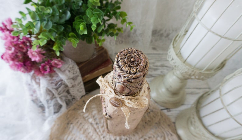 May include: A decorative bottle with a beige and brown finish, featuring a floral design and a lace ribbon. The bottle is sitting on a hessian surface.