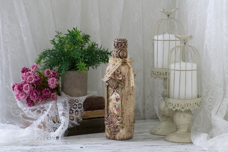 May include: A decorative glass bottle with a beige and brown paint finish, intricate details, and a lace ribbon. The bottle is on a white surface with a lace tablecloth, a potted plant, and two white candles in birdcage holders.