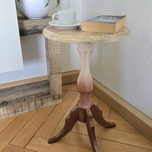 May include: A small, round, wooden side table with a white and brown ombre finish. The table has a single pedestal base with three legs and a round top. A white cup and saucer and a book are on the table.