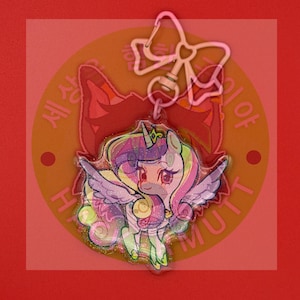 May include: A clear acrylic keychain featuring a cartoon unicorn with a glitter finish, a purple and yellow mane, and wings. The charm is attached to a light pink metal clasp shaped like a bow. The unicorn has large eyes and a small horn.