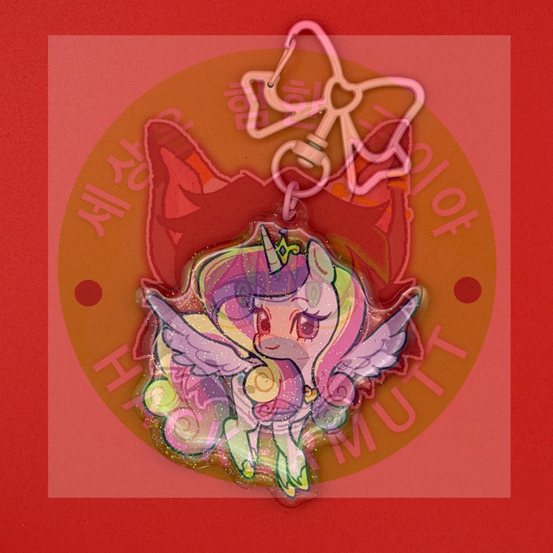 May include: A clear acrylic keychain featuring a cartoon unicorn with a glitter finish, a purple and yellow mane, and wings. The charm is attached to a light pink metal clasp shaped like a bow. The unicorn has large eyes and a small horn.