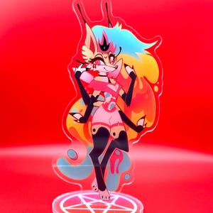 May include: An acrylic standee featuring a stylised anthropomorphic creature with four arms, long ears, and vibrant rainbow-coloured hair. The character wears a pink top and shorts with black limb markings, standing on a circular base with a white pentagram.