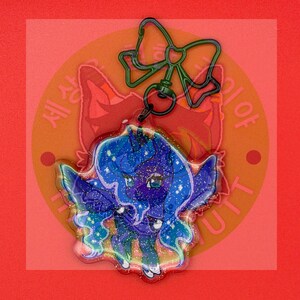 May include: A glittery acrylic keychain featuring a dark blue pony character with a flowing mane and wings, attached to a dark green metal bow-shaped clasp. The character has large eyes and a starry, cosmic pattern throughout its body and hair.