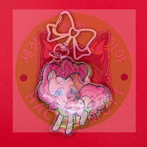 May include: A clear acrylic keychain featuring a cartoon pony with a pink mane and tail, accented with gold glitter. The keychain includes a pink bow-shaped clasp at the top. The design is layered over a circular background with Korean text.