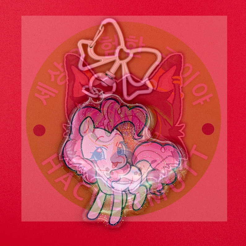 May include: A clear acrylic keychain featuring a cartoon pony with a pink mane and tail, accented with gold glitter. The keychain includes a pink bow-shaped clasp at the top. The design is layered over a circular background with Korean text.