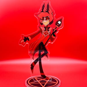 May include: A clear acrylic standee featuring a cartoon character with red hair, wearing a red suit with a black bowtie, and holding a staff with a microphone top. The character stands on a circular base printed with a black and red pentagram design.
