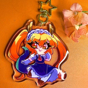 May include: A clear acrylic keychain featuring a cartoon character with long orange hair, wearing a blue and white maid outfit. The character has red eyes and a yellow and orange tail.