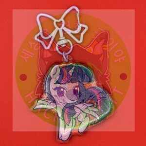 May include: A clear acrylic keychain featuring a glittery, cartoon-style unicorn character with a purple and blue mane. The charm is attached to a white, bow-shaped metal clasp. The character has large eyes and a stylized, colorful appearance.