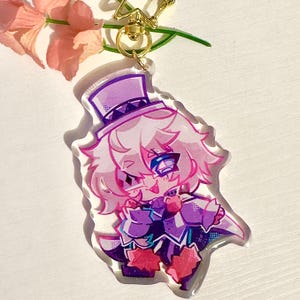 May include: A purple and pink anime character keychain with a white top hat and a mischievous expression. The character is wearing a purple and white outfit with a red heart design.