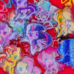 May include: A collection of glittery acrylic charms featuring various cartoon ponies with colourful manes and tails. Each charm displays a unique character with large expressive eyes, set against a vibrant red background with scattered light reflections.