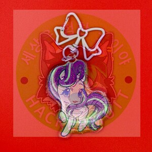 May include: A glittery acrylic keychain featuring a cartoon unicorn with a purple and mint green mane. The charm is attached to a light purple bow-shaped metal clasp. The unicorn has large eyes and a stylized appearance with a sparkling finish.