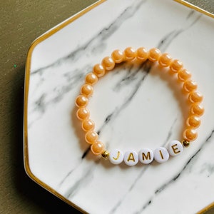 May include: A peach-colored beaded bracelet with the name "JAMIE" spelled out in gold letters on white beads.