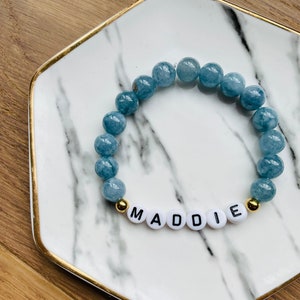 May include: A blue gemstone bracelet with gold beads and white beads spelling out the name "MADDIE".