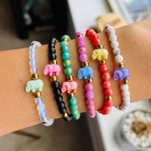 May include: Seven colorful beaded bracelets with small elephant charms. The bracelets are stacked on a person's wrist. The bracelets are made with small beads in various colors, including white, black, turquoise, pink, red, and purple. Each bracelet has a small gold charm in the shape of an elephant.