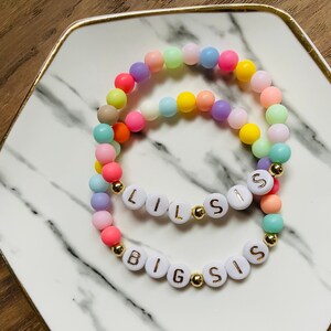 Puede incluir: Dos pulseras de cuentas coloridas con las palabras "Big Sis" y "Lil Sis" escritas en letras blancas con detalles dorados.