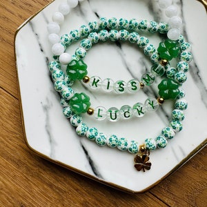 Peut inclure: Trois bracelets superposés avec des breloques et des perles en trèfle vert. La couche supérieure est blanche, la couche intermédiaire a des motifs de trèfles verts et la couche inférieure a des perles transparentes avec les mots "Kiss Me Lucky" écrits. Un charme en trèfle doré pend de la couche inférieure.