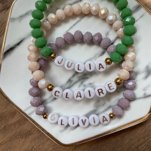 May include: Three beaded bracelets with the names "Julia", "Claire", and "Olivia" spelled out in white beads with gold lettering. The bracelets are layered on a marble surface.