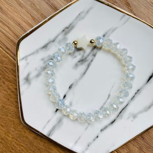 May include: A white star charm bracelet with clear faceted glass beads. The bracelet is on a white marble plate with gold trim.