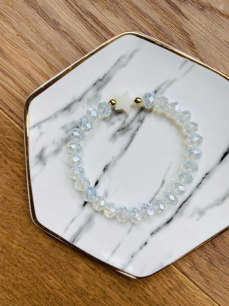 May include: A white star charm bracelet with clear faceted glass beads. The bracelet is on a white marble plate with gold trim.