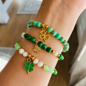May include: Three beaded bracelets in shades of green and white are displayed. Each bracelet features a gold charm, including a four-leaf clover. The bracelets are made with various beads, including round and irregular shapes.
