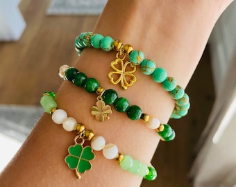 May include: Three beaded bracelets in shades of green and white are displayed. Each bracelet features a gold charm, including a four-leaf clover. The bracelets are made with various beads, including round and irregular shapes.