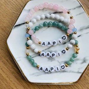 May include: Three layered bracelets with white, pink, and green beads. Each bracelet has a charm with the letters "AAPI" and a heart.