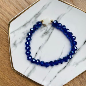 May include: A blue crystal beaded bracelet with a white star charm. The bracelet is on a white marble surface with gold accents.