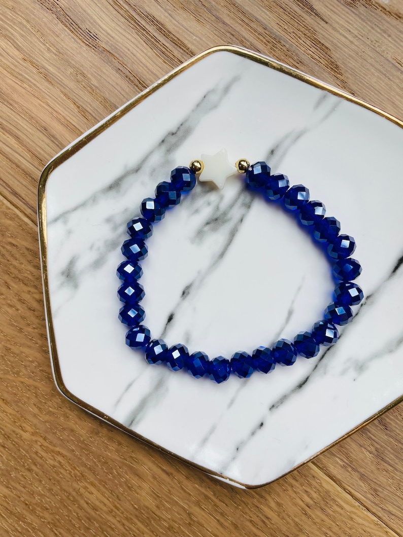 May include: A blue crystal beaded bracelet with a white star charm. The bracelet is on a white marble surface with gold accents.