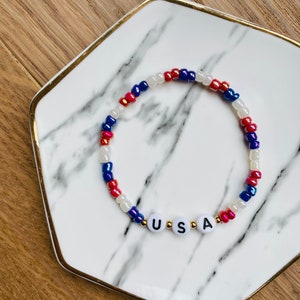 May include: A red, white and blue beaded bracelet with the word "USA" spelled out in white letters on gold beads.