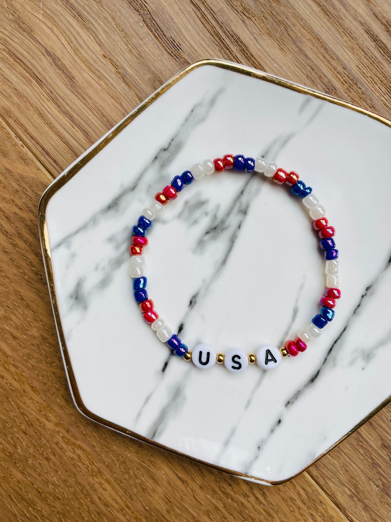 May include: A red, white and blue beaded bracelet with the word "USA" spelled out in white letters on gold beads.