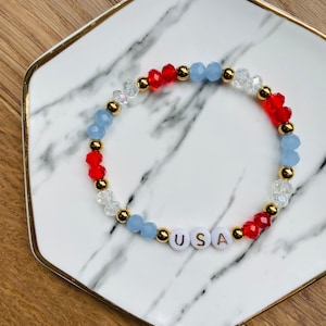 May include: A red, white and blue beaded bracelet with gold accents and the word "USA" spelled out in white beads.