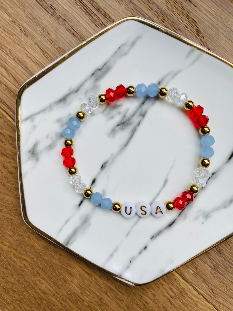 May include: A red, white and blue beaded bracelet with gold accents and the word "USA" spelled out in white beads.
